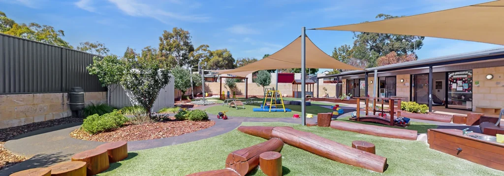 A colorful outdoor playground featuring various play structures, slides, and shade sails. Pathways and tree stumps are present, and the area is surrounded by trees and a fence.