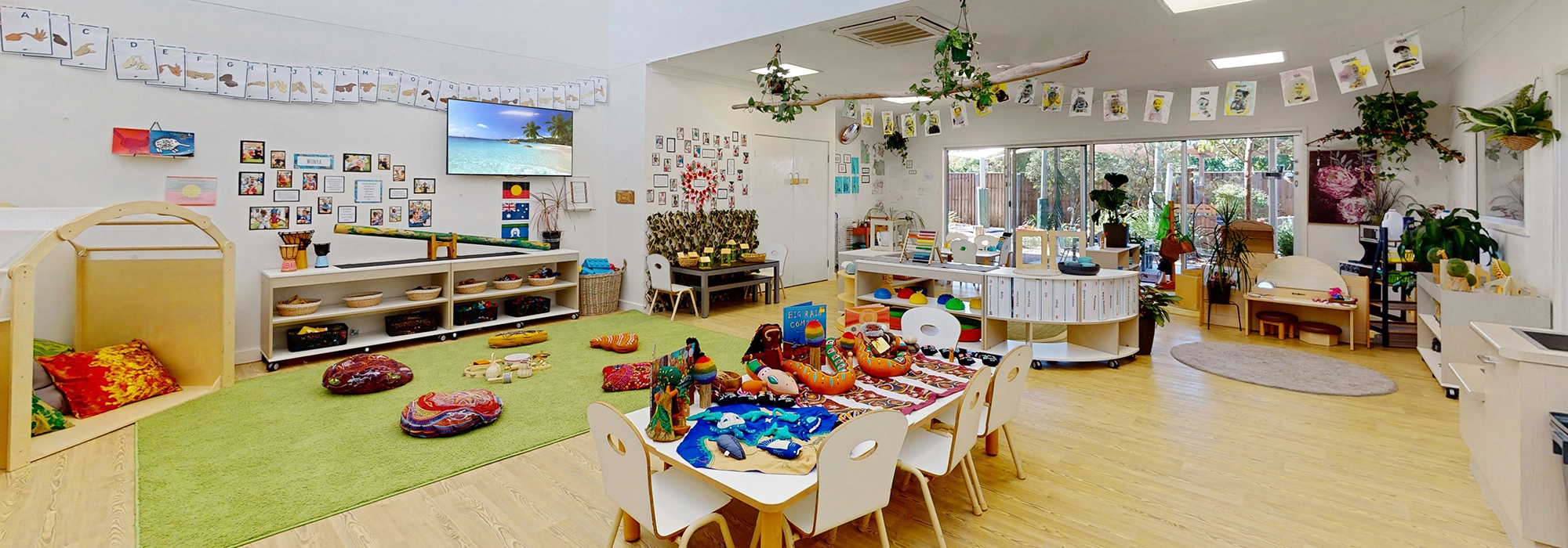 A brightly lit, colorful classroom at Busy Bees Childcare at Burpengary East with various activity areas, including tables, bookshelves and toys. There are plants and children's crafts displayed throughout the room.