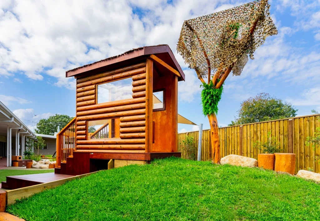 The outdoor playground at Burpengary Daycare centre features a wooden cubby house on a grassy hill, with a wooden tree structure nearby, standing in a fenced yard under a partly cloudy sky.