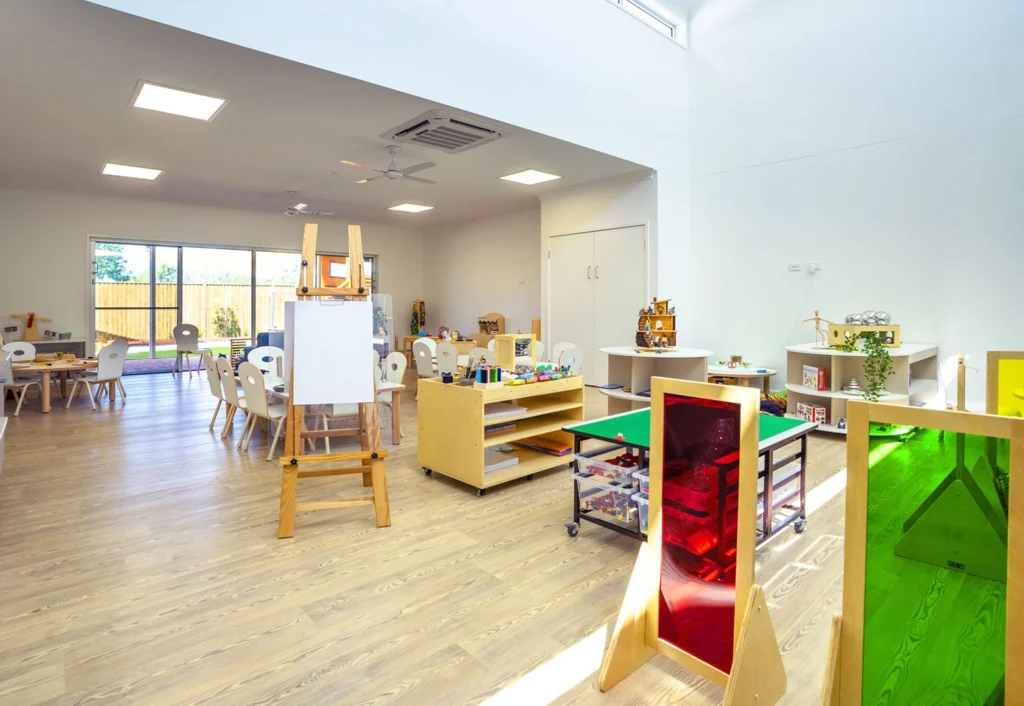 A brightly lit kindergarten classroom with wooden floors, tables and chairs, an easel, storage units, and various educational materials. Large windows allow natural light to enter the room.