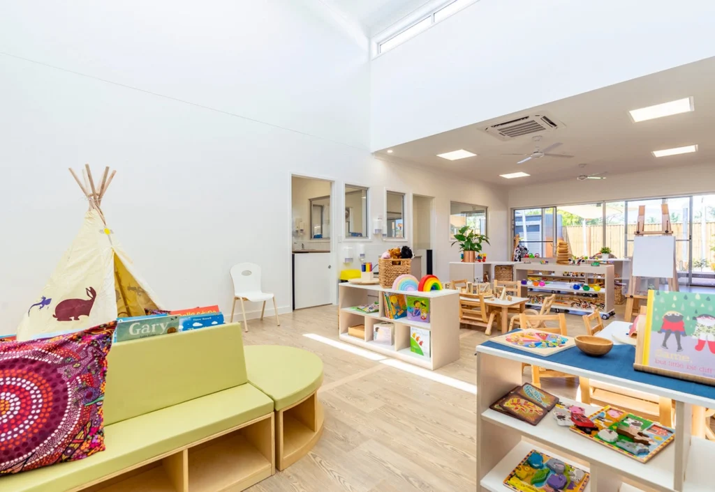 A brightly lit, well-organized children's playroom at Burpengary Childcare with a fabric teepee, activity tables, chairs, bookshelves, and various toys and educational materials.