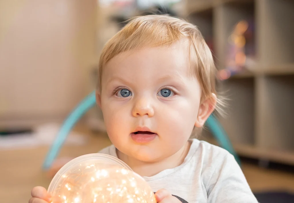 A baby with light hair and blue eyes looking directly at the camera. There are blurred shelves and toys in the background.