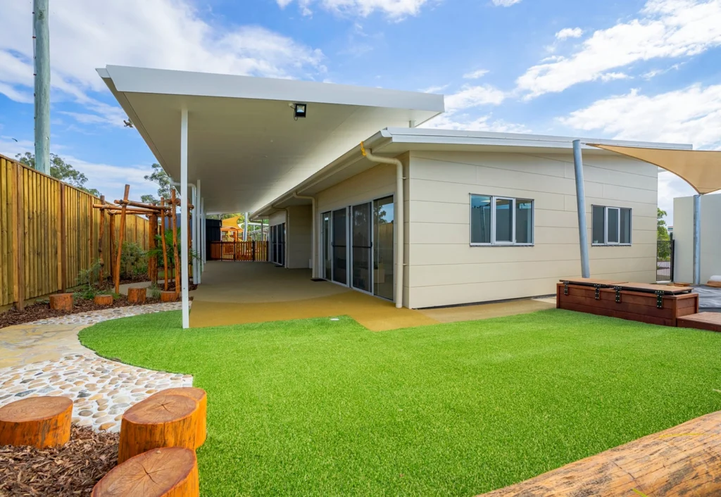 A modern one-story building with large windows, an extended covered patio, artificial grass and wooden stepping logs at Busy Bees Childcare in Burpengary.