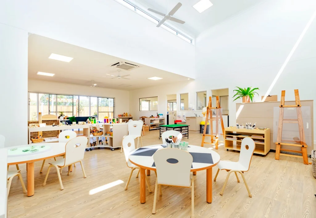 A brightly lit kindergarten classroom at Busy Bees Burpengary East with round tables, chairs, easels, and various educational materials arranged neatly.