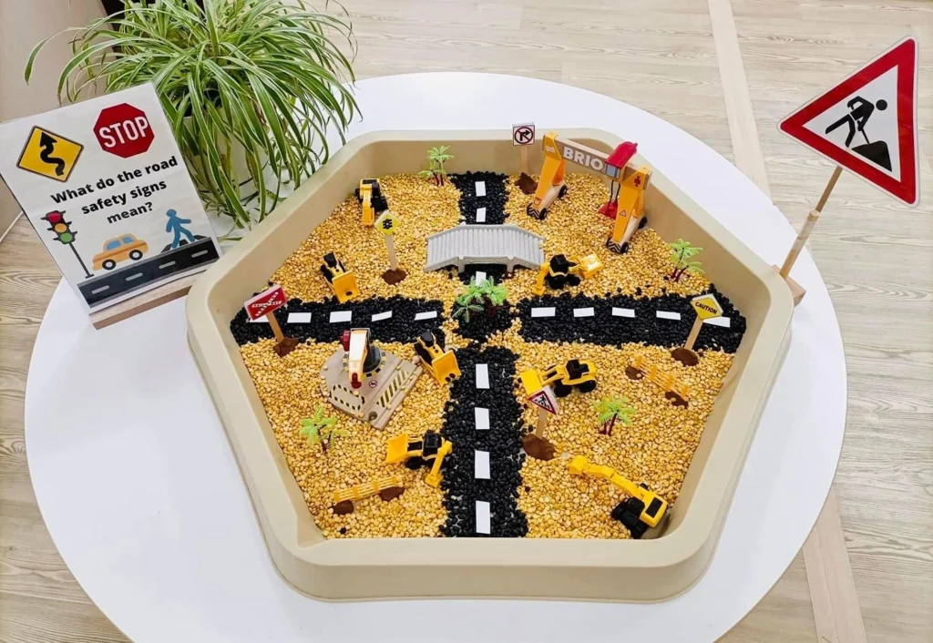 A hexagonal tuff tray displays a miniature construction site with toy vehicles, road signs, and a model road intersection. A signboard on the left asks about the meaning of road safety signs.