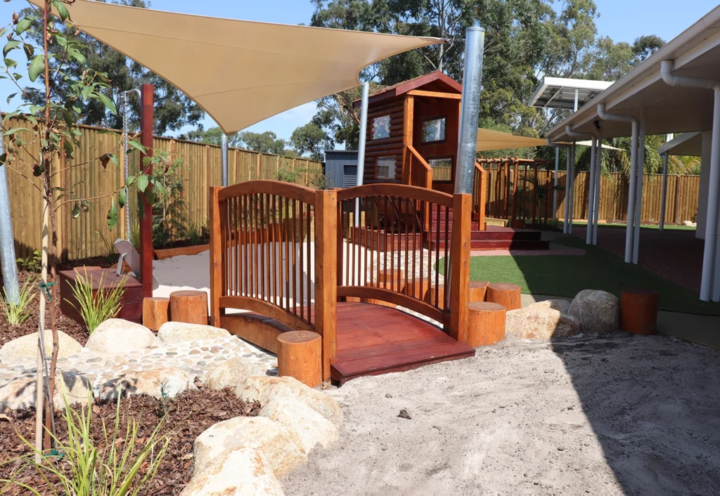 A small wooden bridge leads to a sandpit under shade sails and a wooden playhouse in the background surrounded by a fence and greenery at the Burpengary Daycare.