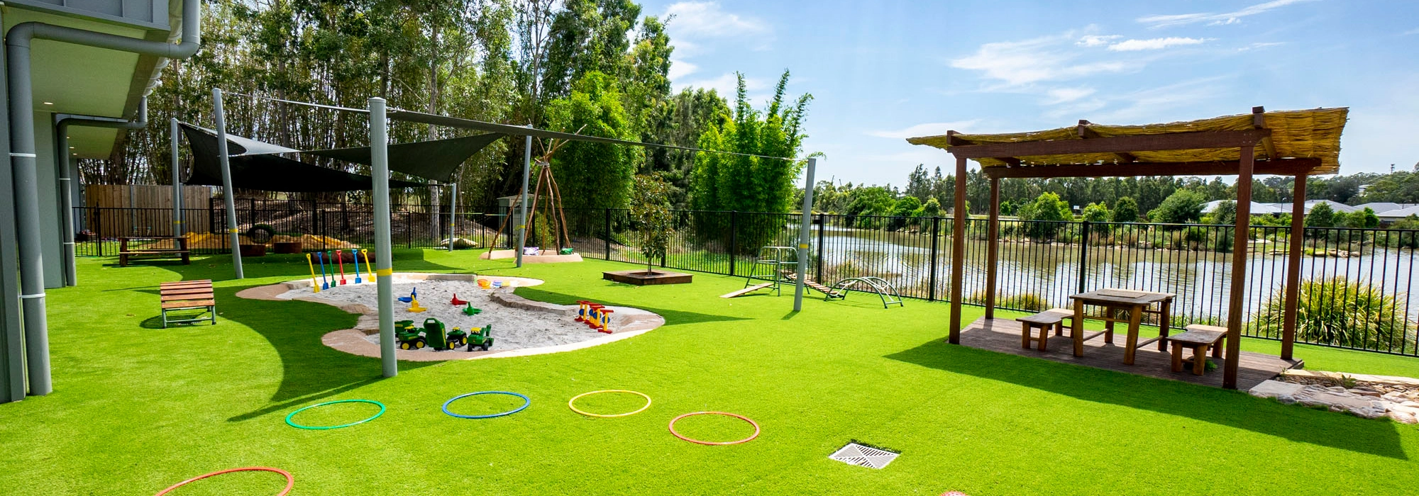 A children's playground with artificial grass, a sandbox, various play equipment, and picnic tables under a wooden canopy, located near a body of water and surrounded by trees.