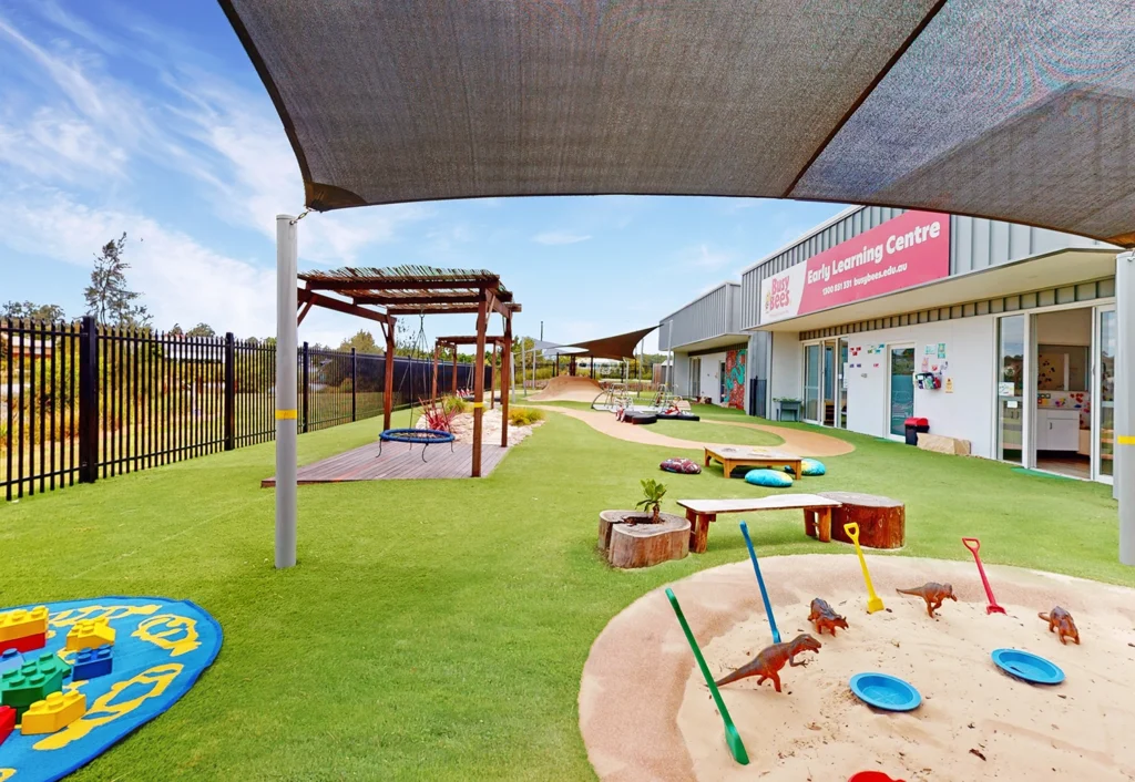 Outdoor playground area at a childcare center with a sandbox, toys, shade structures, and a building labeled "Early Learning Centre" in the background.