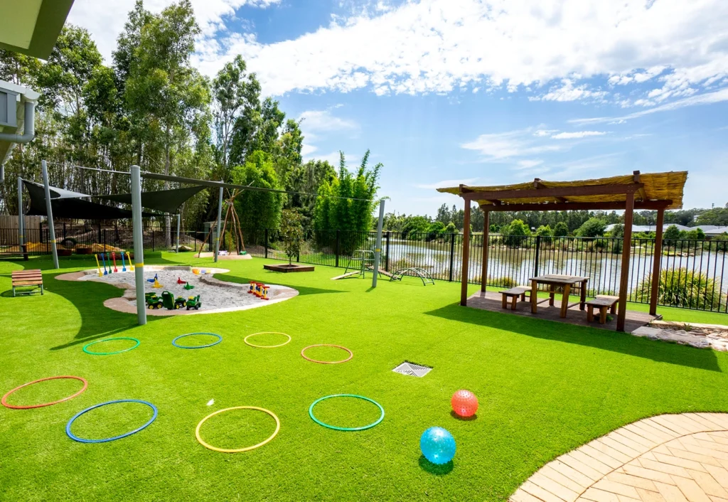 Outdoor playground with green artificial grass, colorful hula hoops, a sandbox, a shaded area, benches, and a small gazebo overlooking a lake and surrounded by trees under a partly cloudy sky.