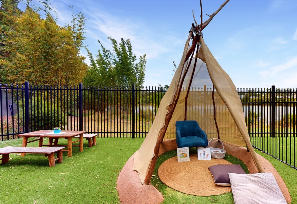 A backyard setup with a teepee-style tent, a blue chair, pillows, and children's books inside. Adjacent is a wooden picnic table on a grass surface, enclosed by a black iron fence.