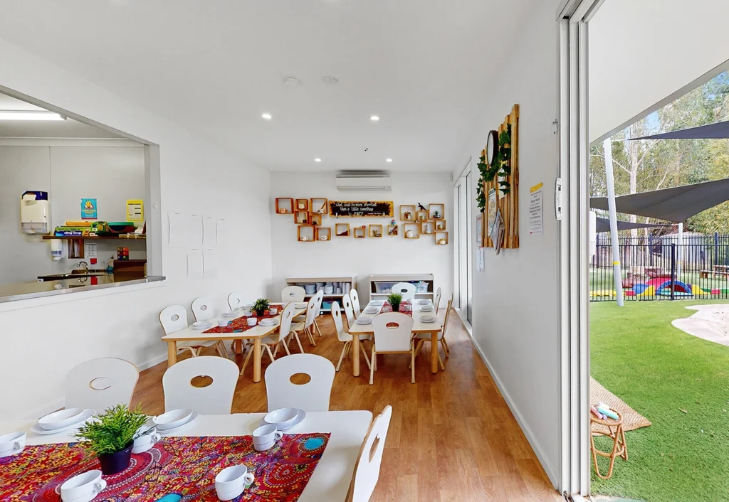 A brightly lit, modern daycare dining room with small tables and chairs set with dishes, colorful tablecloths, and potted plants. An open doorway leads to an outdoor play area with green turf.