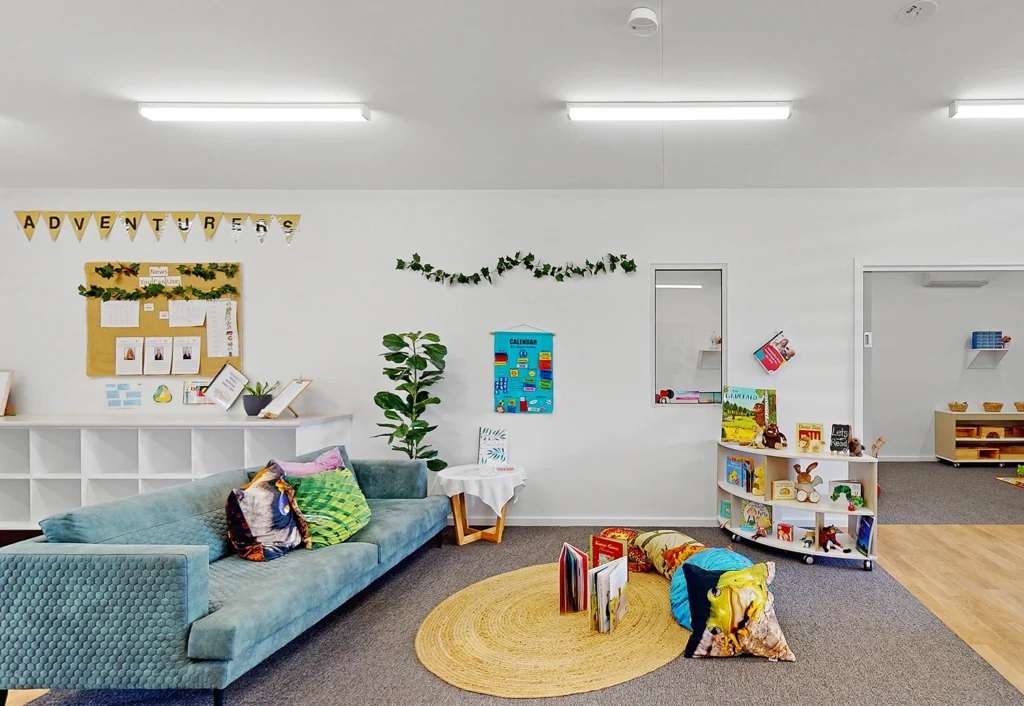 A brightly lit classroom with a blue couch, colorful cushions, a circular rug with books, wall decorations, a bulletin board, potted plants, and shelves with various educational materials.