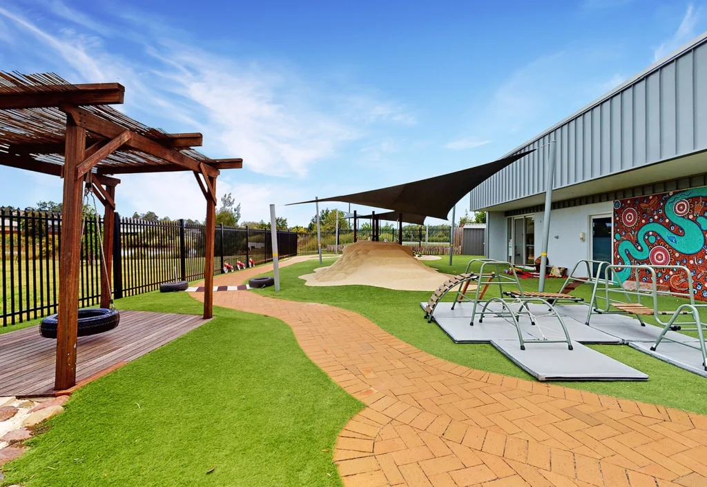 Outdoor play area with synthetic grass, wooden pergola, brick pathway, sandbox with shade sail, climbing frames, and a building with a colorful mural.