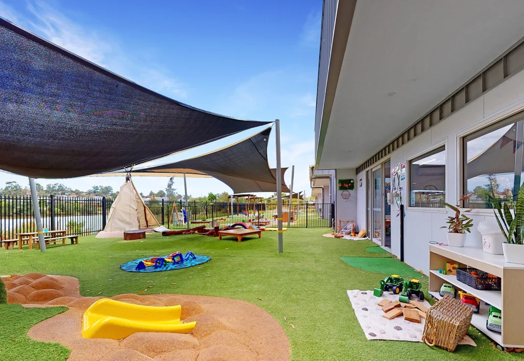 Outdoor play area with artificial grass, a small slide, covered sandpit, and toys. Shade sails provide cover over the space, and a building with windows and a door is on the right.