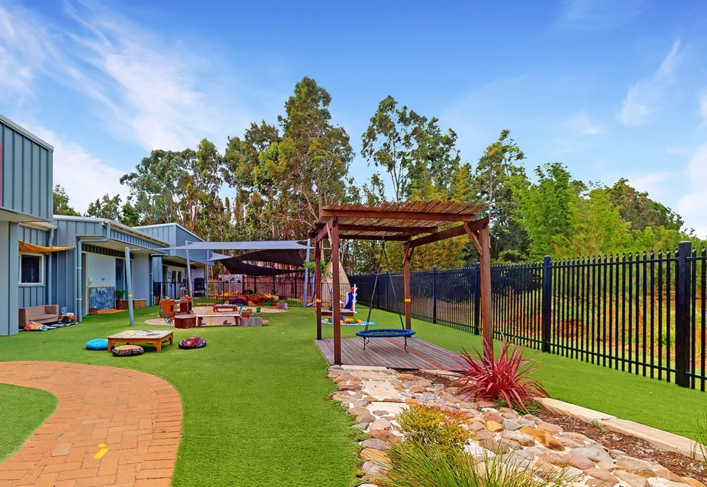 Outdoor playground featuring shade structures, a swing, seating area with cushions, and a grassy area with a stone pathway. Buildings are visible on the left, and tall trees are in the background.