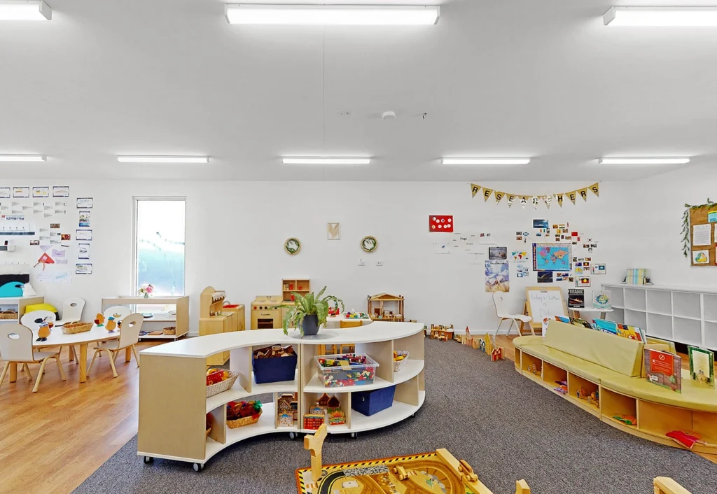 A brightly lit classroom with white walls, low shelves, tables, chairs, educational toys, and a play area. The room is decorated with student artwork and educational posters.