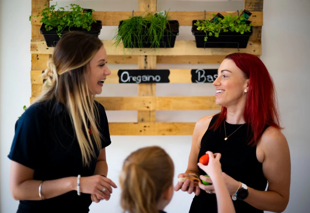 Two women and a child converse in front of a wooden wall-mounted herb planter holding oregano and basil.