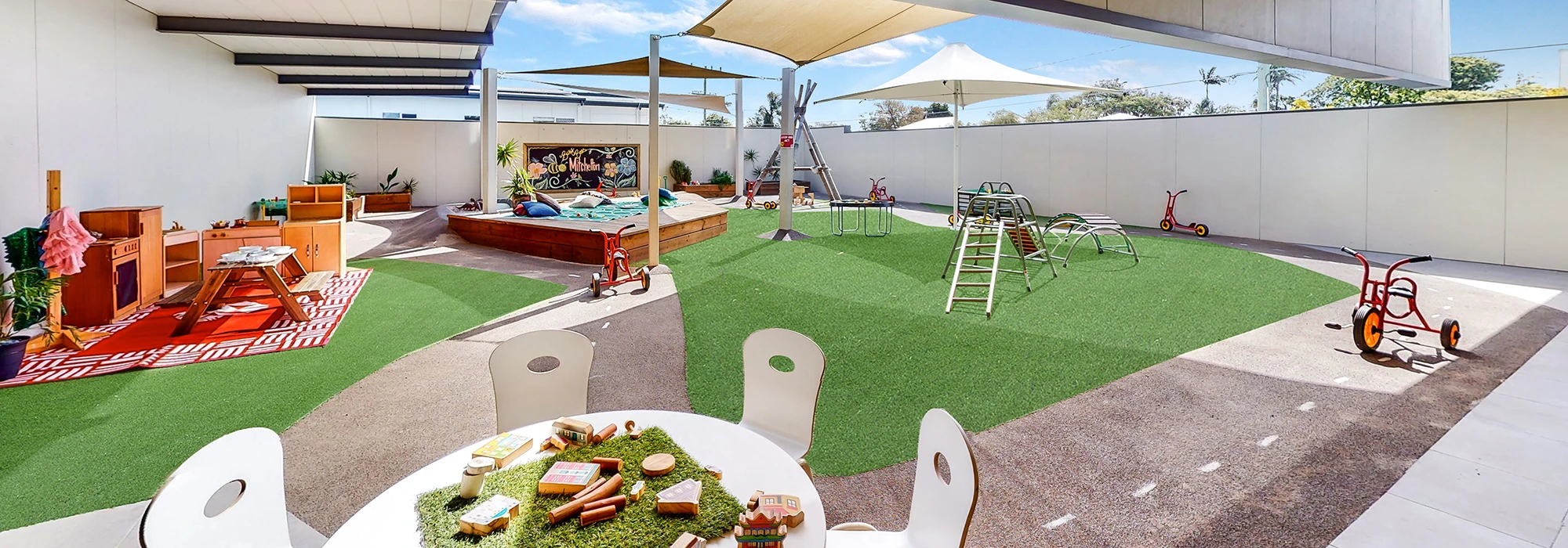 A children's outdoor play area with artificial grass, various playground equipment, tricycles, a sandbox, and a shaded seating area with chairs and a table.