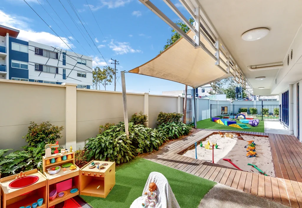 Outdoor play area with a sandbox, toys, and a shaded section. The space is bordered by a wall and features a mix of artificial grass and wooden decking. Nearby buildings are visible in the background.