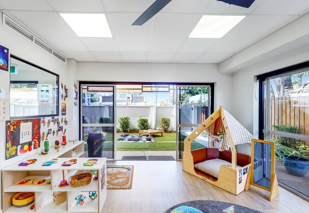 A brightly lit children's playroom with crafts displayed on shelves, a cozy reading nook, and view of an outdoor play area with artificial grass and toys.