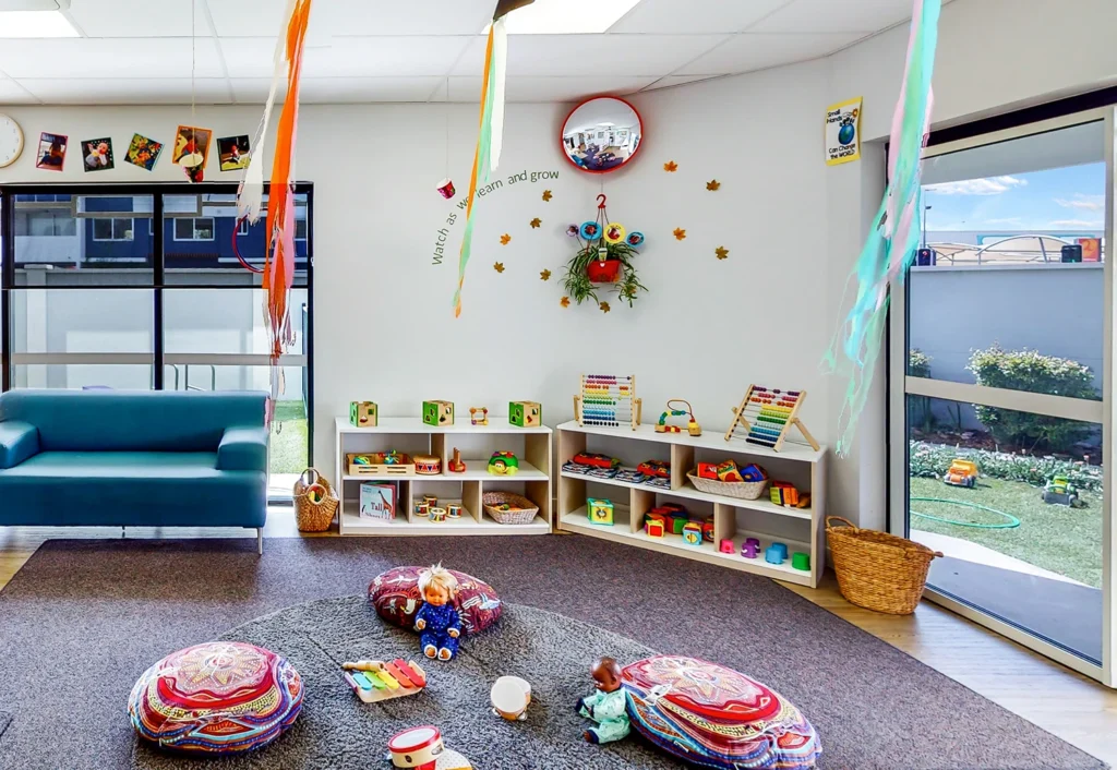 A colorful children's playroom with cushions, toys, and wall decorations. Shelves hold various toys, and a round mirror is on the wall. Windows let in natural light and a door leads outside.