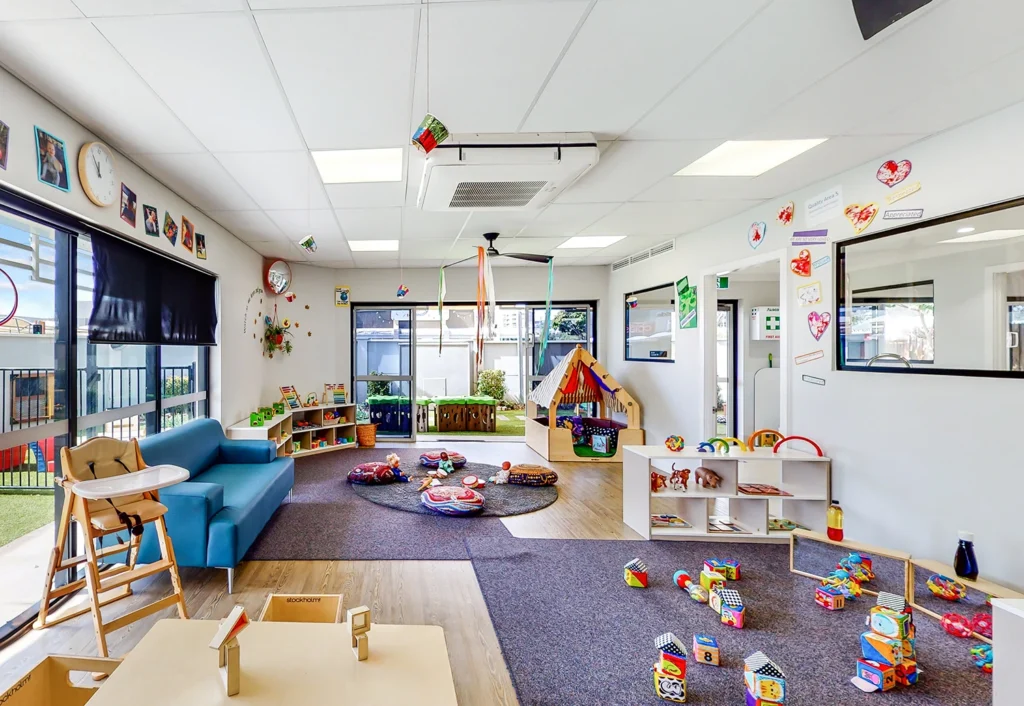 A brightly lit daycare room filled with various toys, books, and play structures, including a tent and a blue couch. Windows allow natural light in, and colorful decorations adorn the walls.