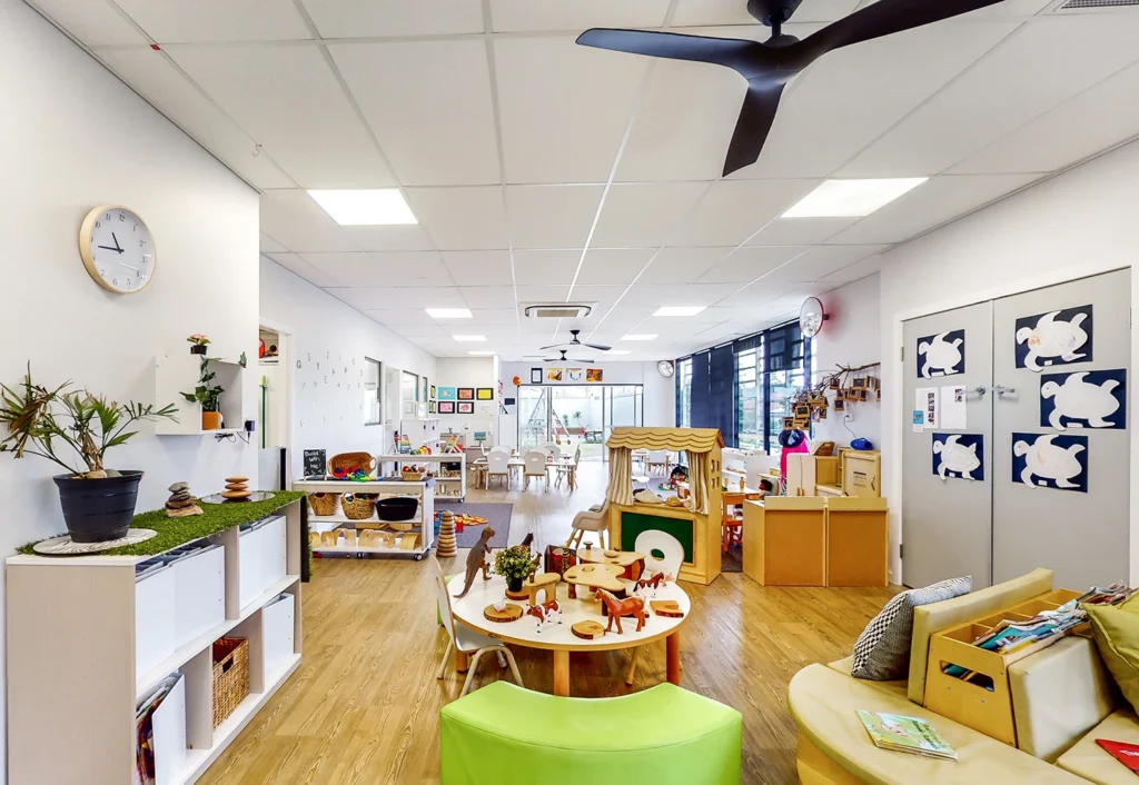 A brightly lit children's playroom with tables, toys, plants, and educational materials neatly arranged. The room features a wooden floor, white walls, a ceiling fan, and large windows.