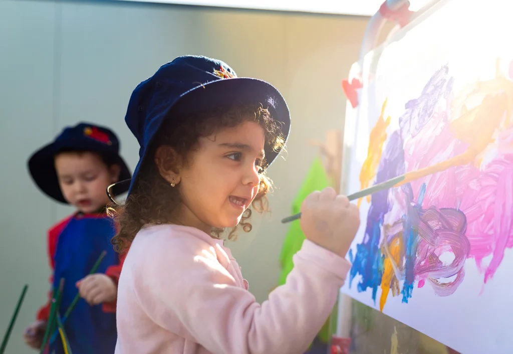 Two young children wearing hats are painting on easels outdoors. One child is smiling while painting, and the other is focused on their work in the background.