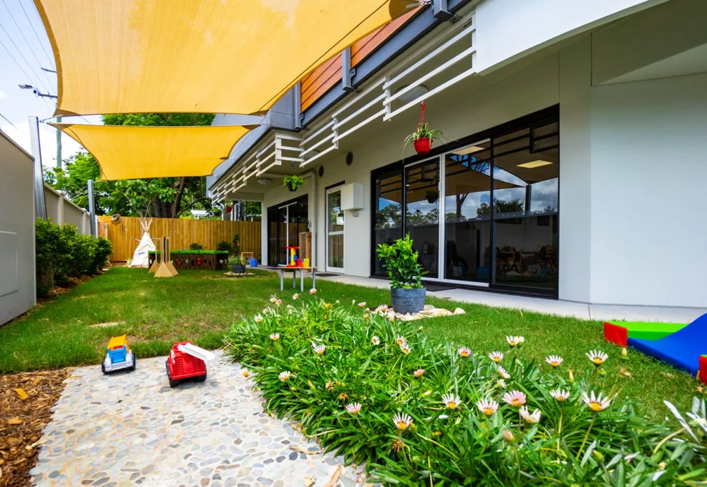 Outdoor play area with toy trucks, a teepee, and various play structures. A building with large windows and shade sails is in the background, surrounded by greenery and flowers.