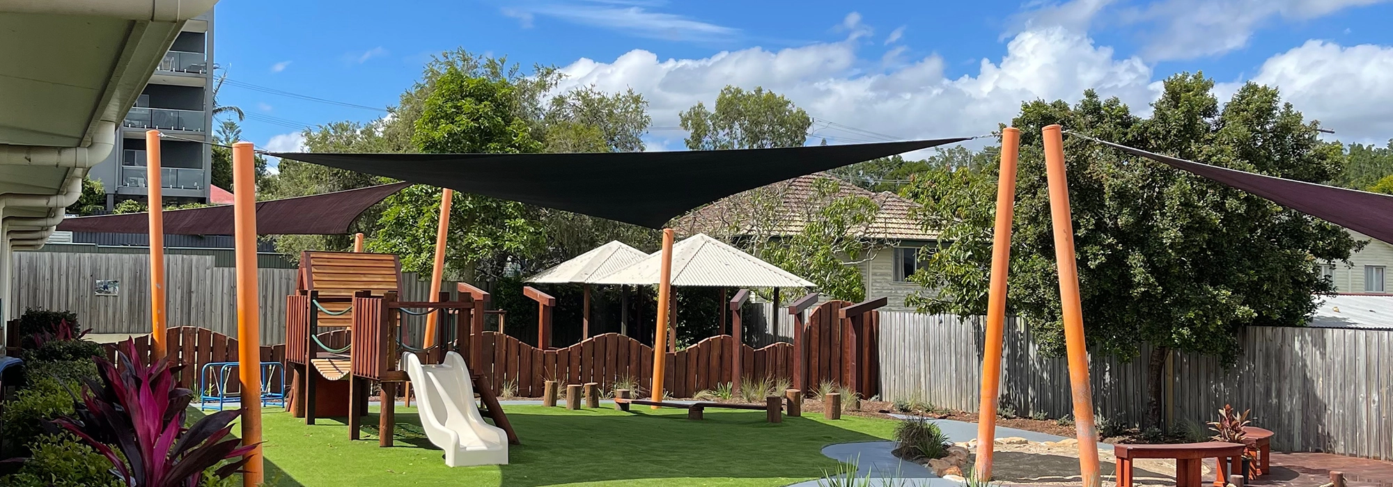 Outdoor playground at Busy Bees Beenleigh childcare with various play structures, including a slide and wooden climbing structure, under shade sails and surrounded by greenery and a wooden fence on a sunny day.