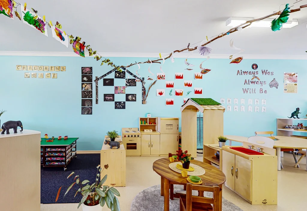 A brightly decorated classroom at Beenleigh daycare featuring a play kitchen, a small table, and various educational displays on the walls, including children's artwork and a tree branch with hanging crafts.