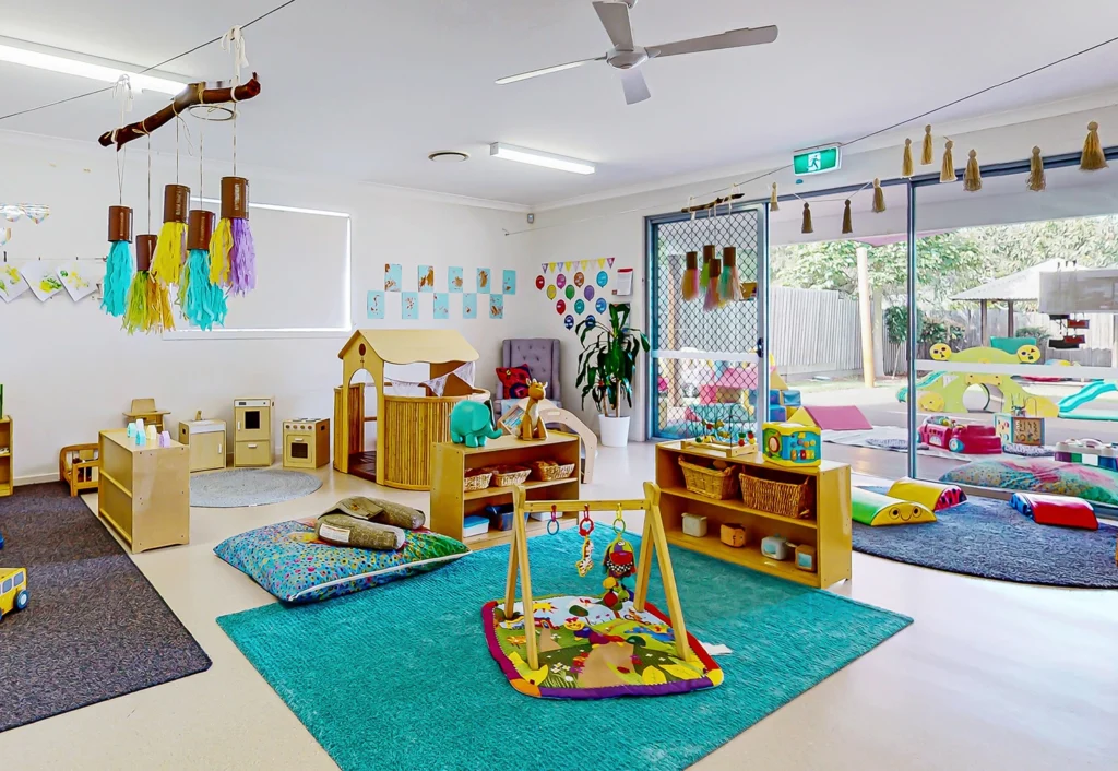 A colorful daycare playroom for babies with various toys, activity centers, and cozy resting areas. The room is well-lit with natural light from large windows and has a door leading to an outdoor play area.