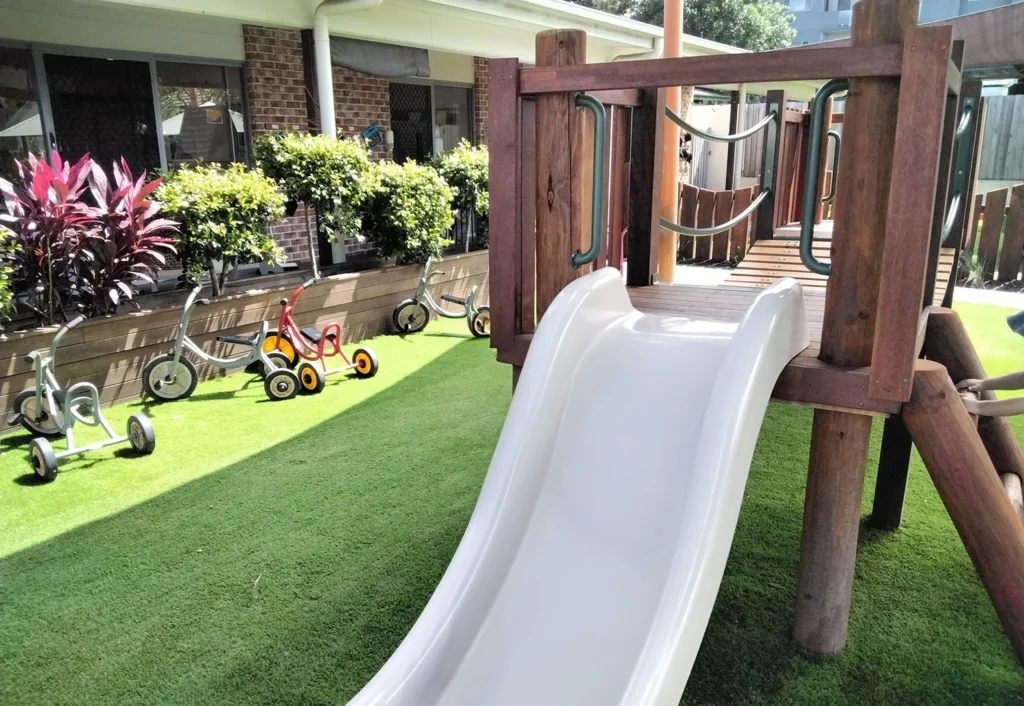 Outdoor playground at Beenleigh Day Care with a slide, artificial grass, tricycles, and a wooden play structure near a building with garden plants.