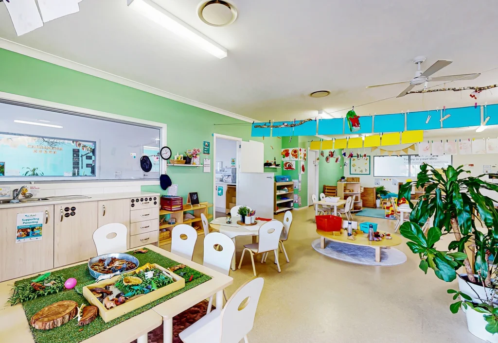 A brightly lit classroom with colorful decorations, various play areas, small tables and chairs, and educational materials neatly arranged at Beenleigh childcare.