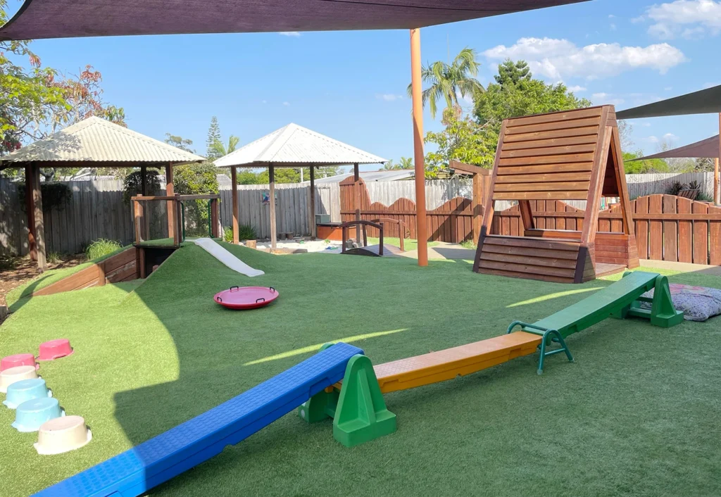 Outdoor playground at Beenleigh child care centre featuring a wooden trinagle cubby house, a slide, bridge, shaded areas, and play equipment under a sunny sky.