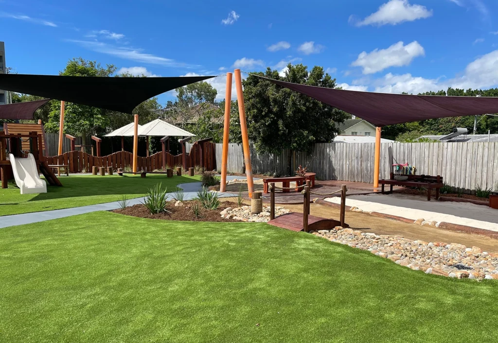 Outdoor playground at Beenleigh Early Learning Centre with artificial grass, shade sails, a slide, wooden structures, and a sandbox surrounded by landscaping. Fence and trees in the background, under a partly cloudy sky.