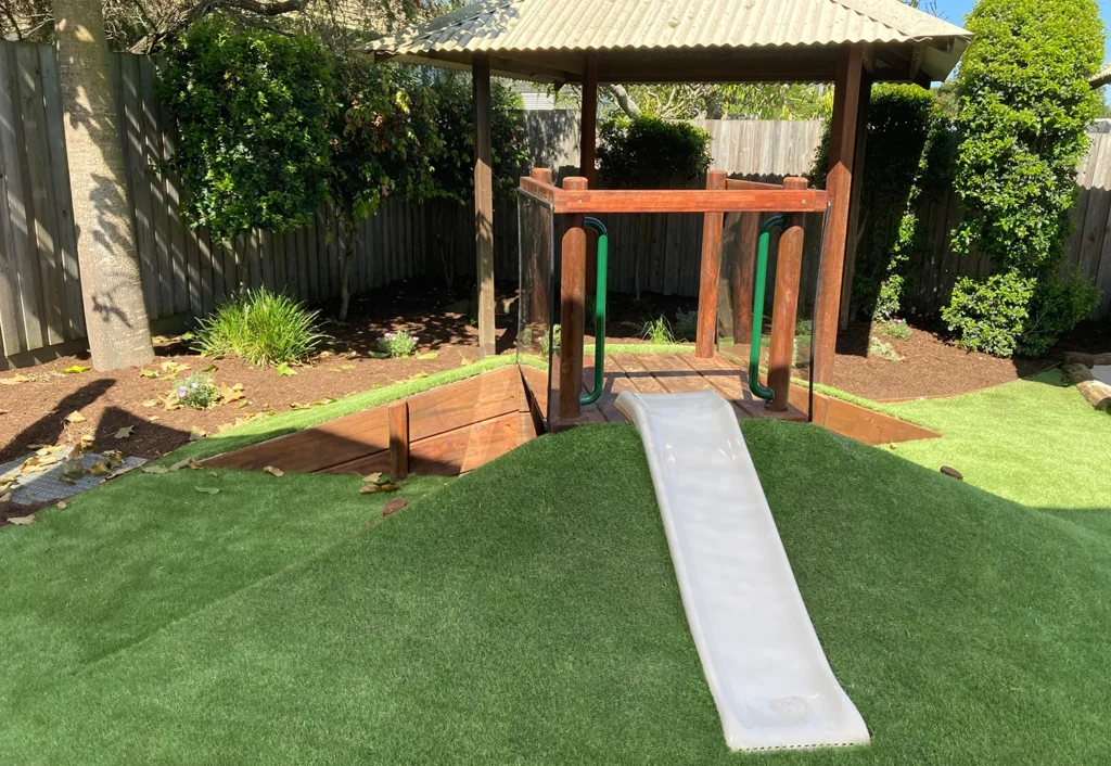 Nursery playground at a day care in Beenleigh featuring a wooden play structure with a small white slide siting on a grassy mound.