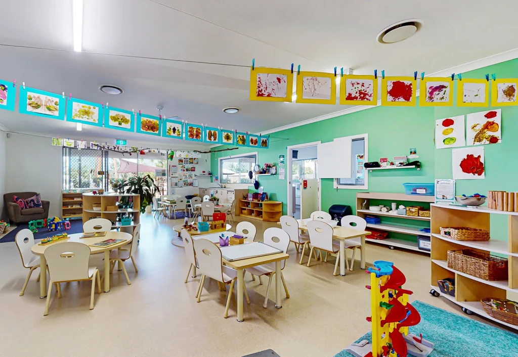 A brightly colored classroom at a Beenleigh Day Care with tables and chairs, art supplies, and children's artwork hanging from a string. Shelves hold educational materials and toys. Walls are painted a vibrant green.