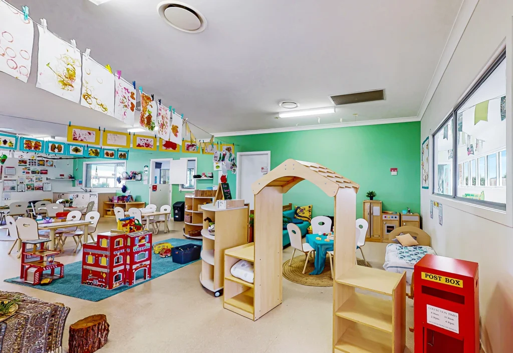 A colorful and organized kindergarten classroom at Busy Bees Beenleigh with children's artwork on display, small tables and chairs, wooden shelves, a playhouse, a red postbox, and various educational materials.