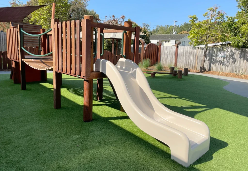 A children's playground at Beenleigh Day Care featuring a wooden play structure with an arched bridge and a white dual slide.