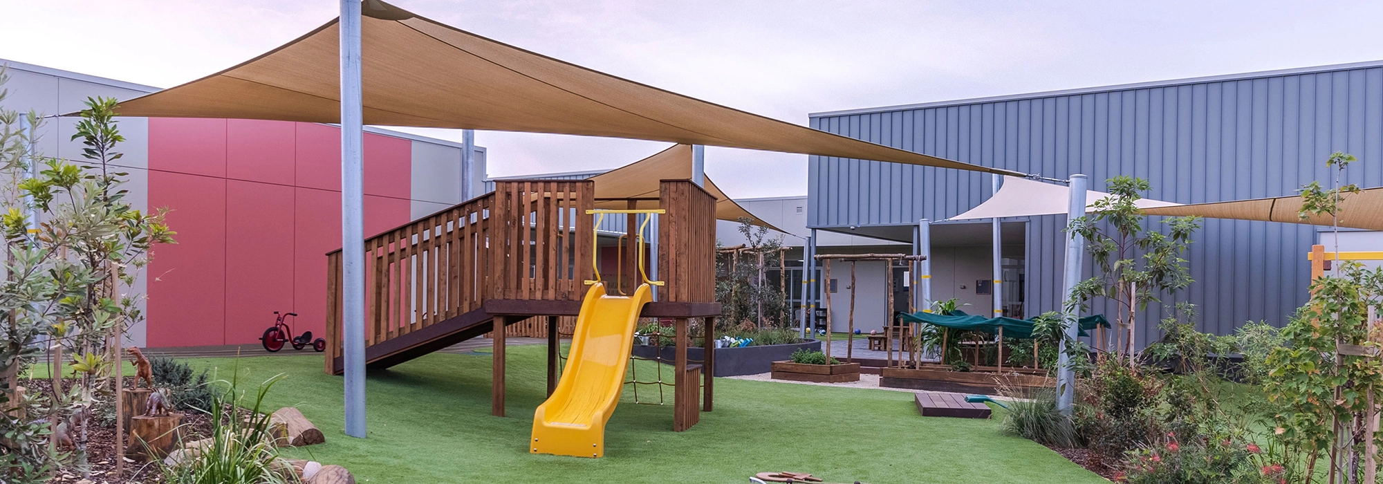 Outdoor playground at Cameron Park childcare with a wooden climbing structure, a yellow slide, and shade sails. The playground has artificial grass, surrounding plants, and nearby modern buildings.