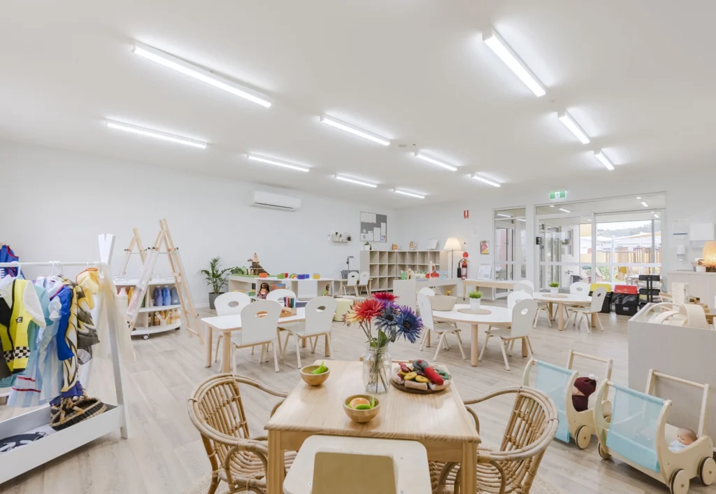 A brightly lit, modern preschool classroom with tables, chairs, bookshelves, and various educational toys and activities. The room has an organized layout with ample space for children to play.