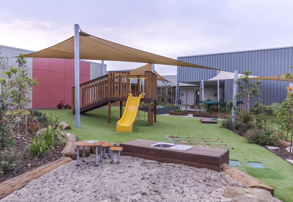 A day care playground with a yellow slide, wooden climbing structure, sandpit, and a grassy outdoor space near modern early learning buildings at Cameron Grove.