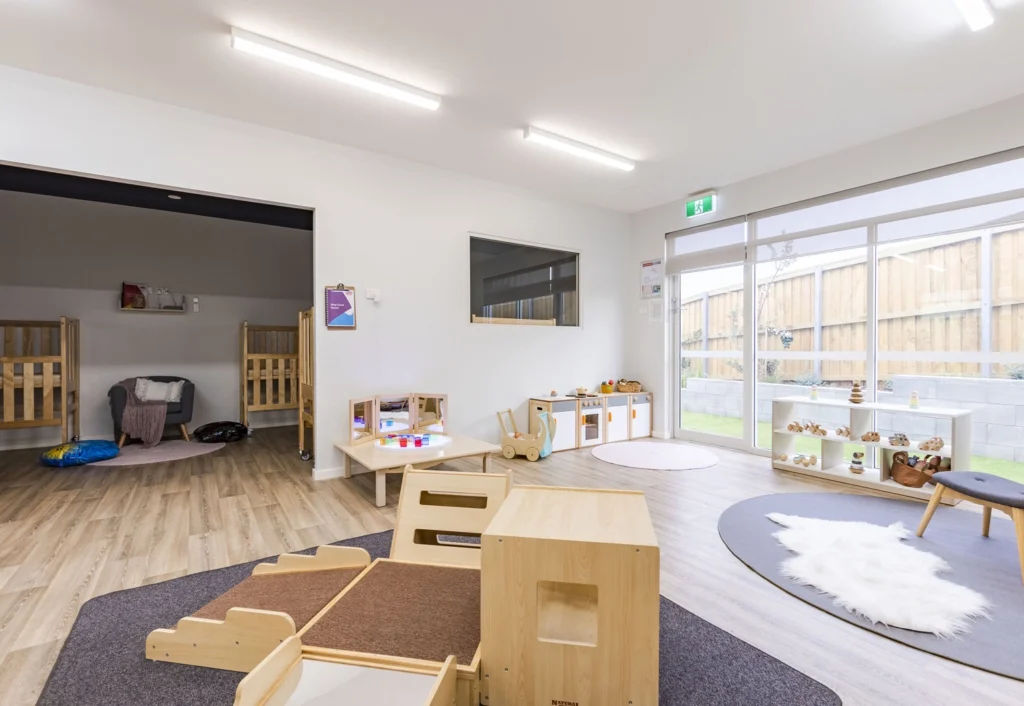 A brightly lit nursery day care room at Busy Bees Cameron Grove with play areas, toys, and small wooden furniture. There's a rug on the floor, a cot room in the background, and large windows letting in natural light.