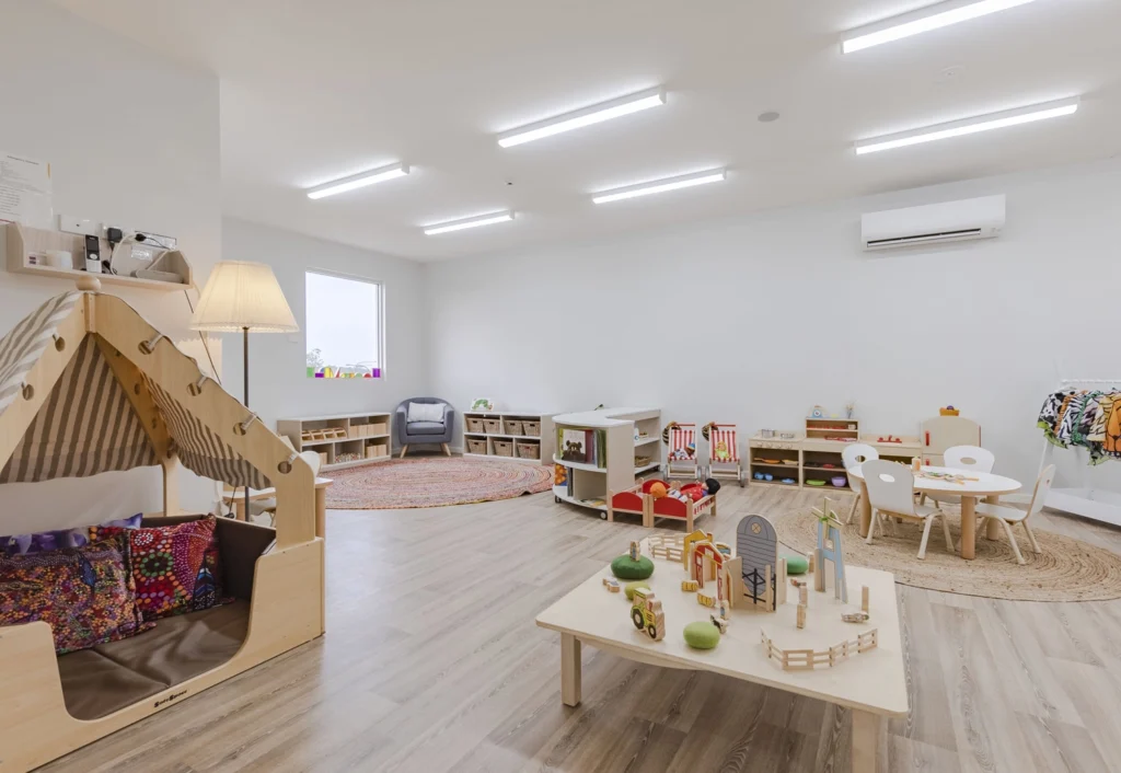 A spacious, well-lit children's playroom at Busy Bees Cameron Park with various toys and furniture, including a reading tent, small tables and chairs, and a pretend play kitchen.