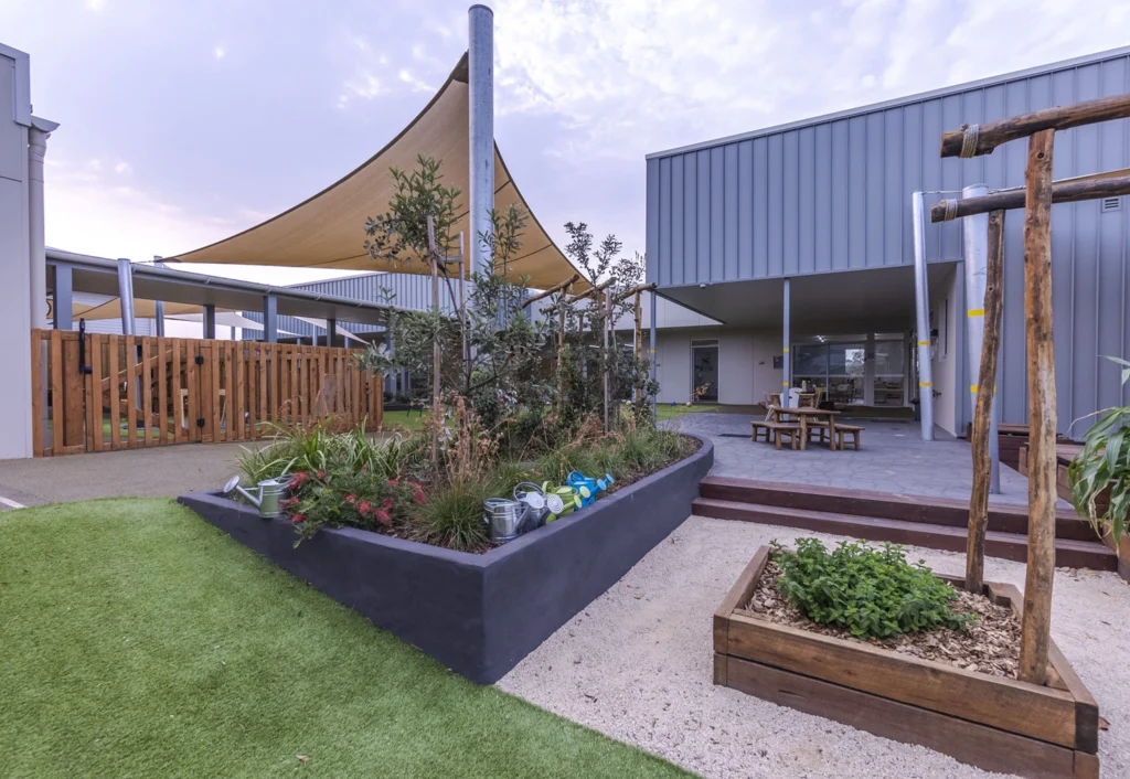 A modern outdoor area at a Cameron Park day care centre with raised garden beds, a shaded seating area, and a wooden fence in the background. The space is surrounded by contemporary architecture and landscaping.