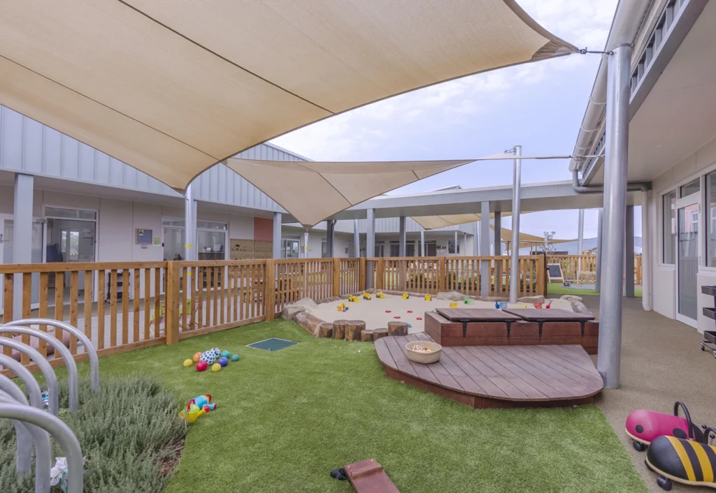 Cameron Park daycare playground or babies featuring a sandpit, various colorful toys and large shade canopies. The area is surrounded by a wooden fence with an adjacent building in the background.