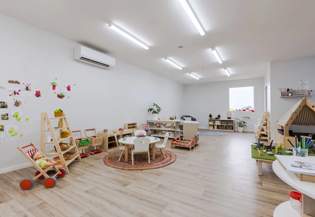 A brightly lit, spacious classroom at Busy Bees Day Care in Cameron Park. The room has a pretend play kitchen, a round table, shelves with toys, and children's artwork decorating the wall.