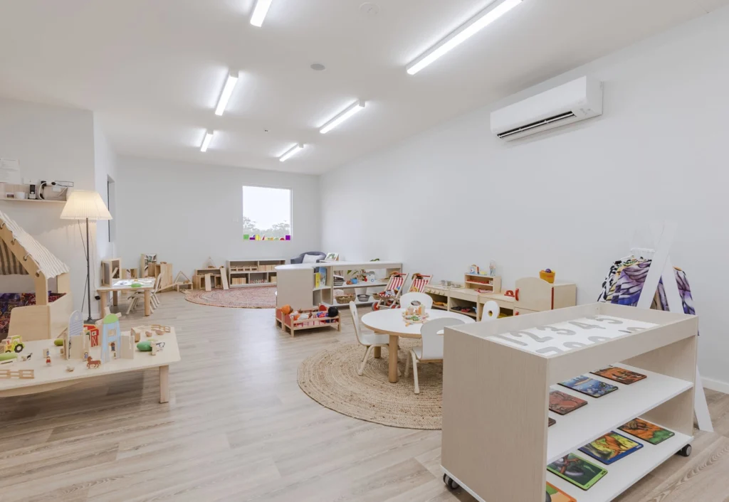 A brightly lit and neatly organised play room at Busy Bees Cameron Grove, featuring wooden flooring, white walls, small tables, chairs, and shelves with toys, and educational materials.