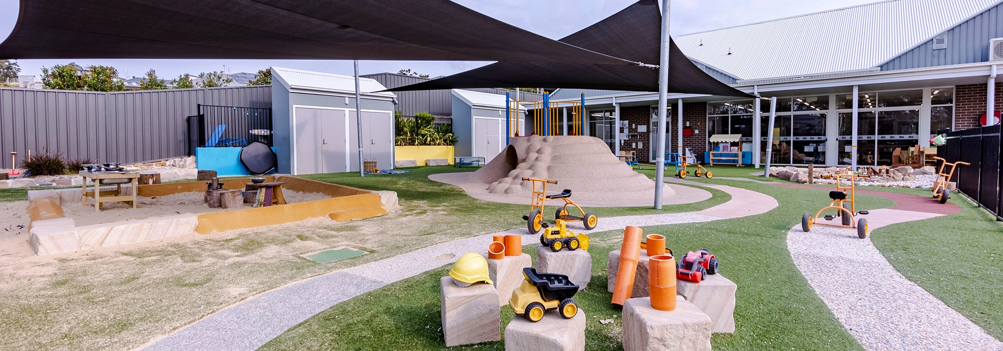 Outdoor playground area featuring a sandpit, small play structures, and various toy vehicles. The area is covered with shade sails and surrounded by a building with large windows.